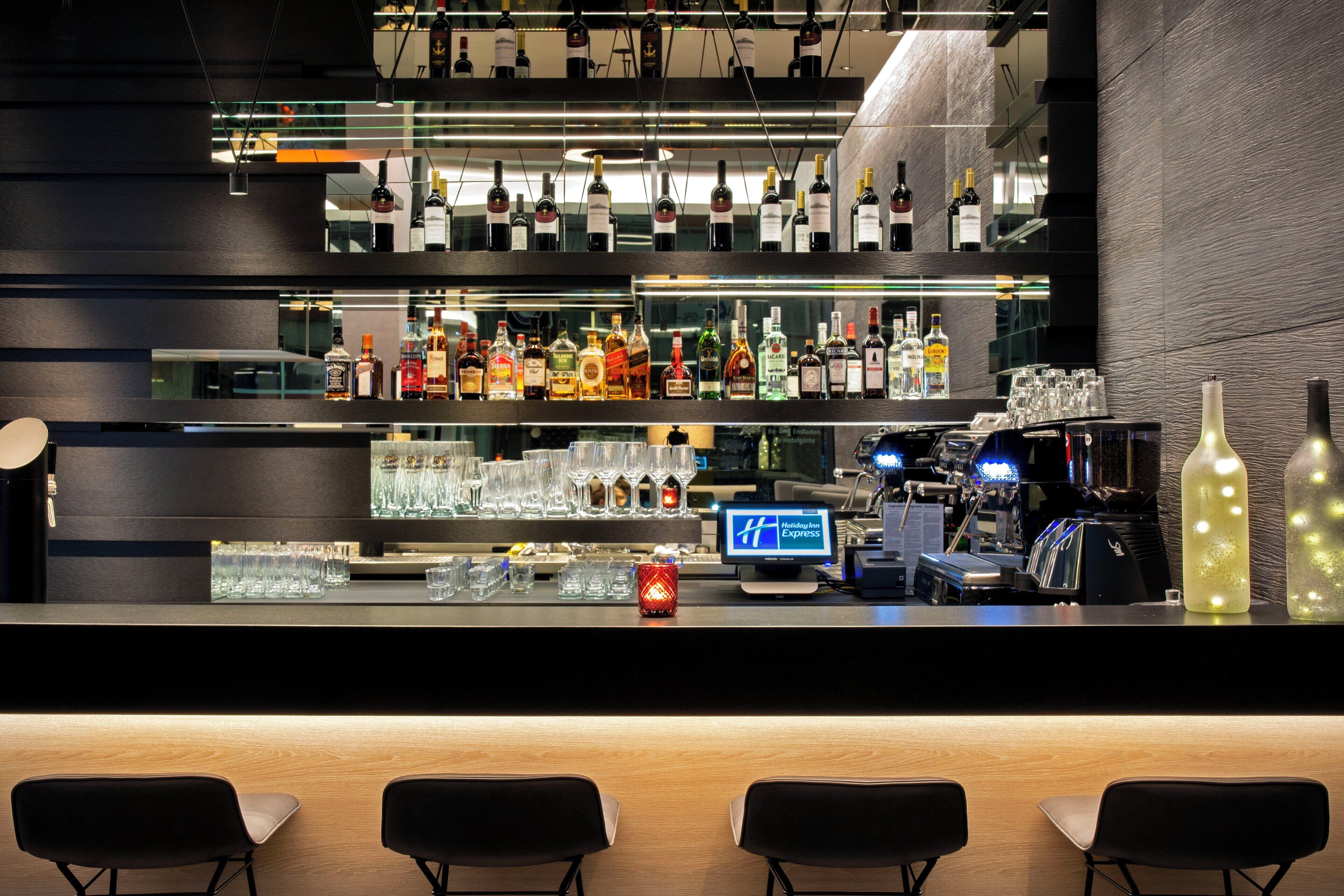 A bar area with multiple shelves of liquor bottles, a digital screen displaying a logo, and bar stools.