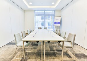 Une salle de conférence avec une longue table, des chaises, un tableau blanc et une fenêtre équipée de stores.
