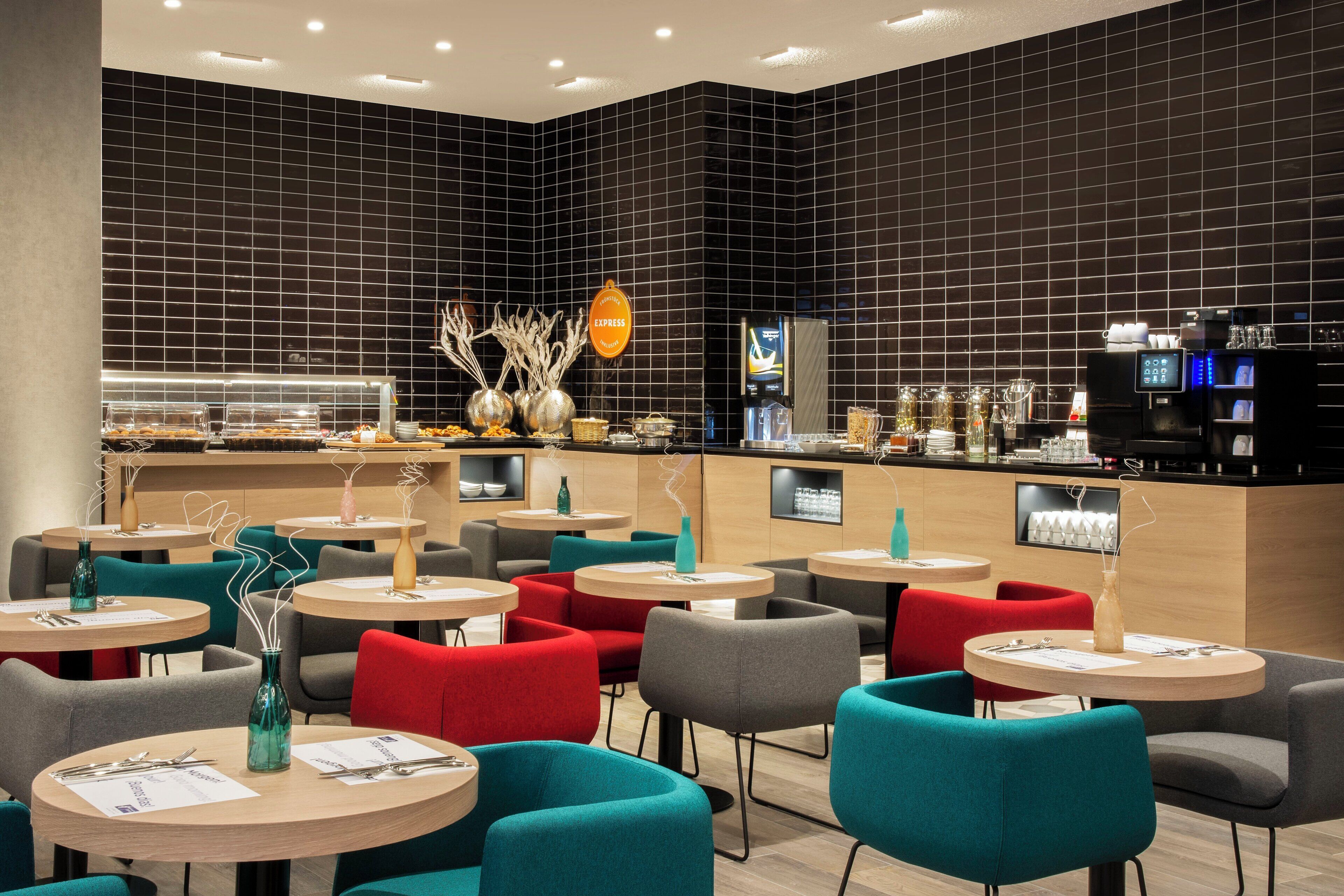 A modern cafe with round tables, teal and red chairs, and a black-tiled wall with a coffee machine and a menu display.