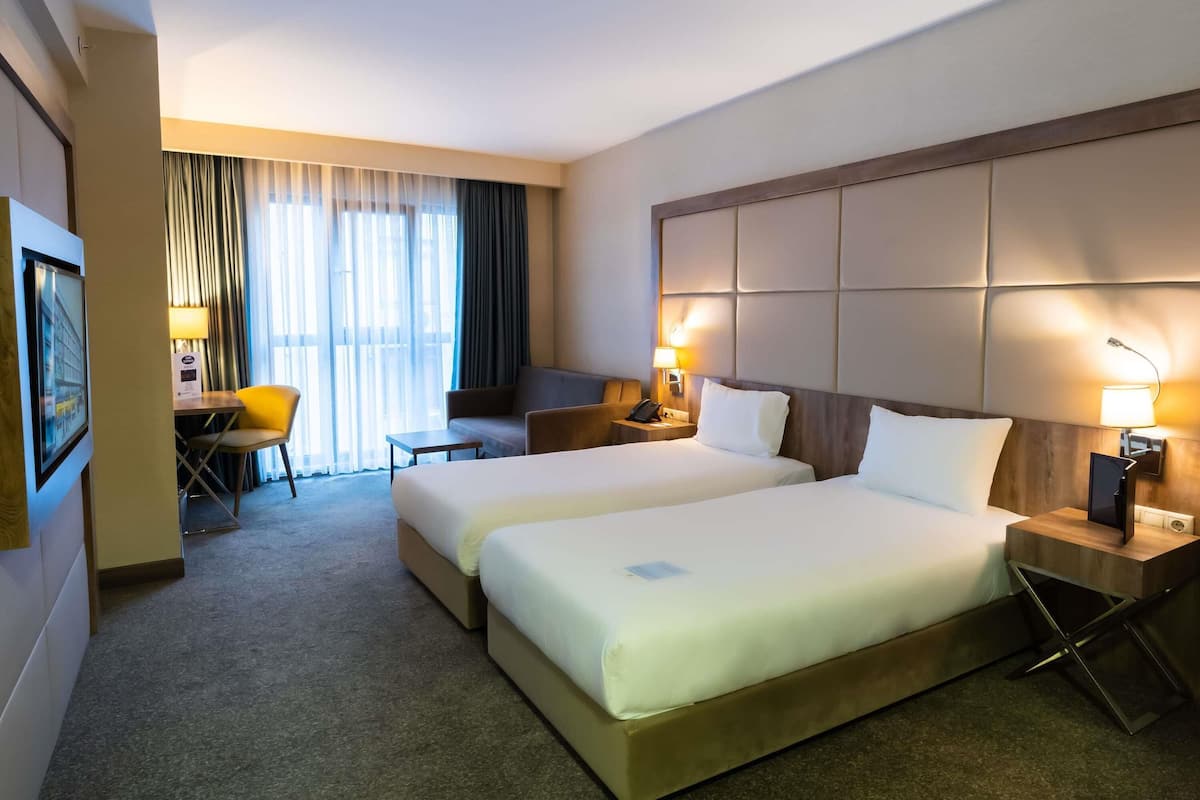 Habitación de hotel con dos camas, un escritorio, una silla y un ventanal con cortinas.