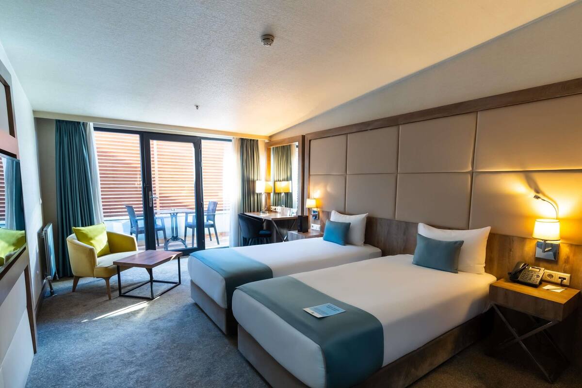 Habitación de hotel con dos camas, un escritorio, una silla, una mesita y un balcón con cortinas.