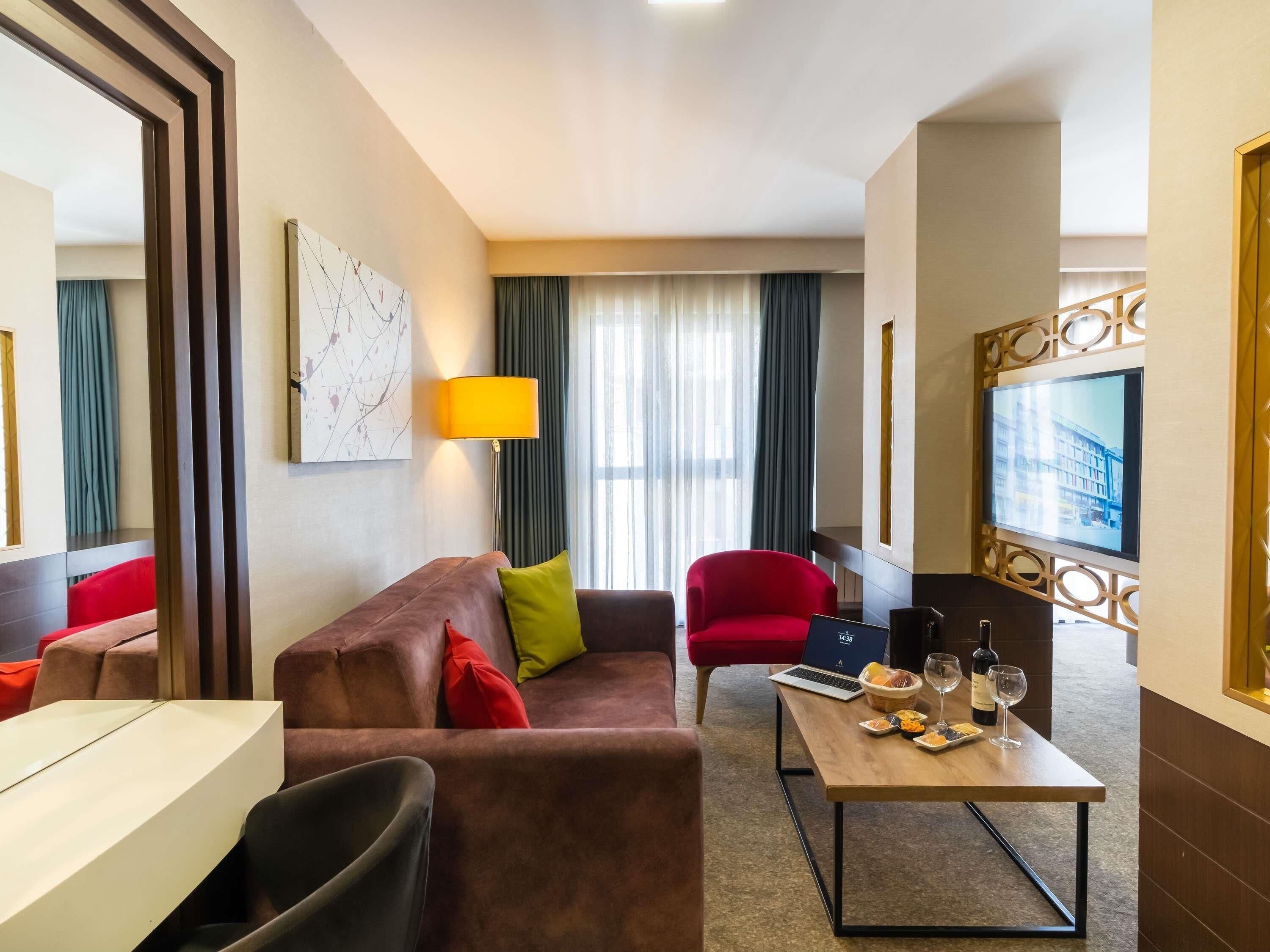 A hotel room with a brown sofa, a red armchair, a wooden coffee table with food and drinks, a TV, and a lamp.