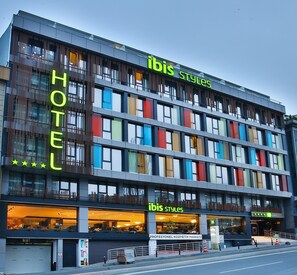 A hotel with a modern design, featuring multiple floors and large windows. The hotel has a sign that reads "ibis Styles" and another sign indicating "PROFESYONEL KOZMETIK MARKETİ".