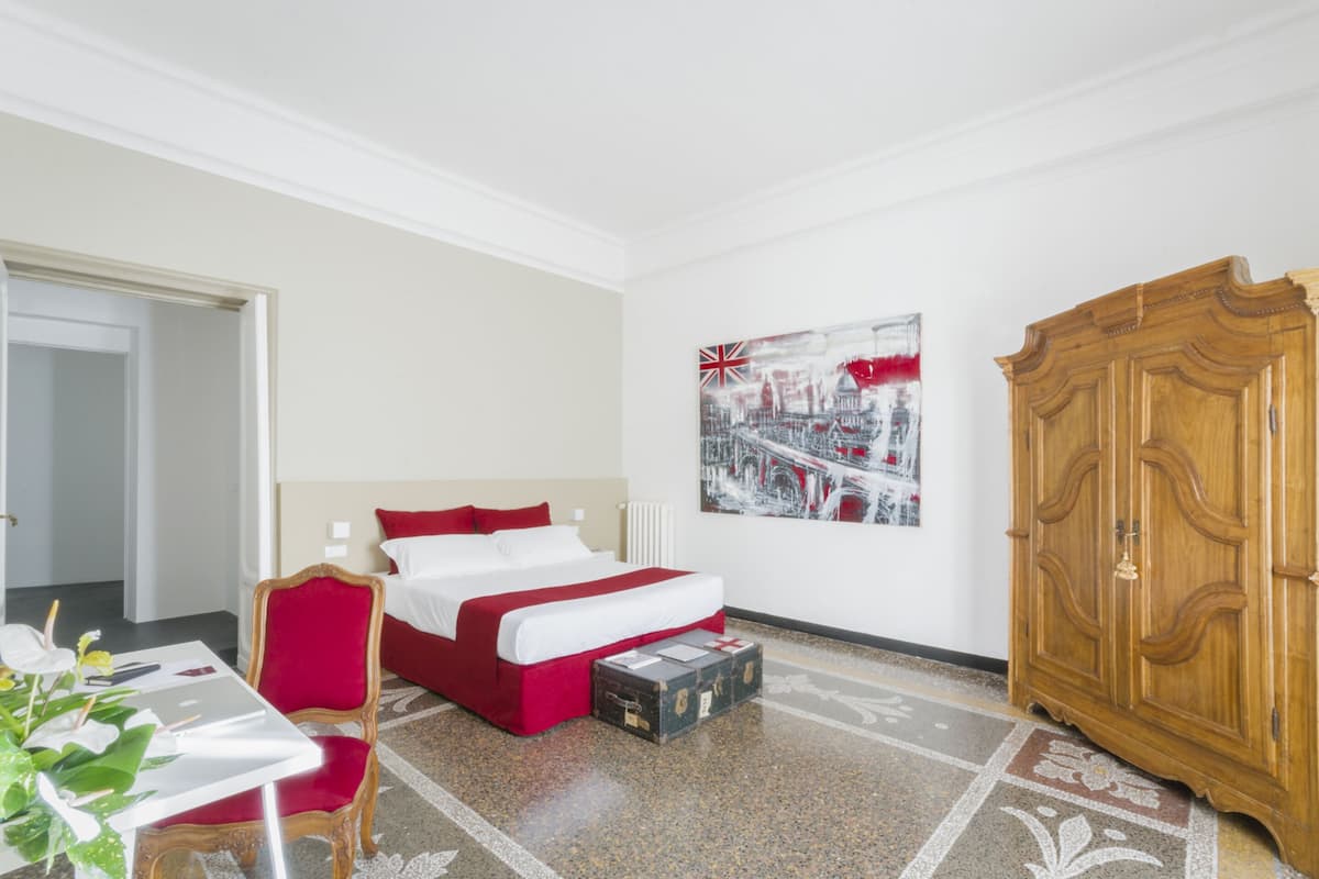 A hotel room with a bed, a wooden wardrobe, a painting on the wall, and a table with a chair.