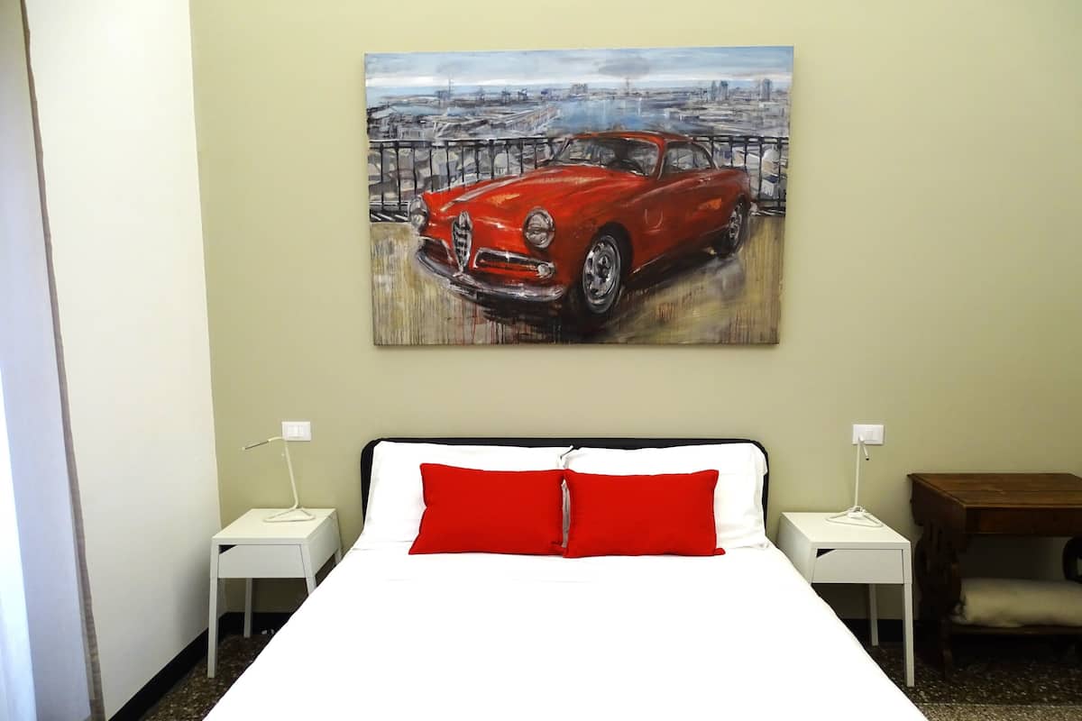 A hotel room with a bed, a red pillow, a nightstand, a lamp, and a painting of a car on the wall.