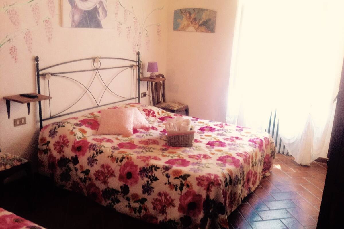 Ein Schlafzimmer mit einem Metallbettgestell, floralem Bettbezug, einem Nachttisch mit Lampe und einem an der Wand montierten Regal.