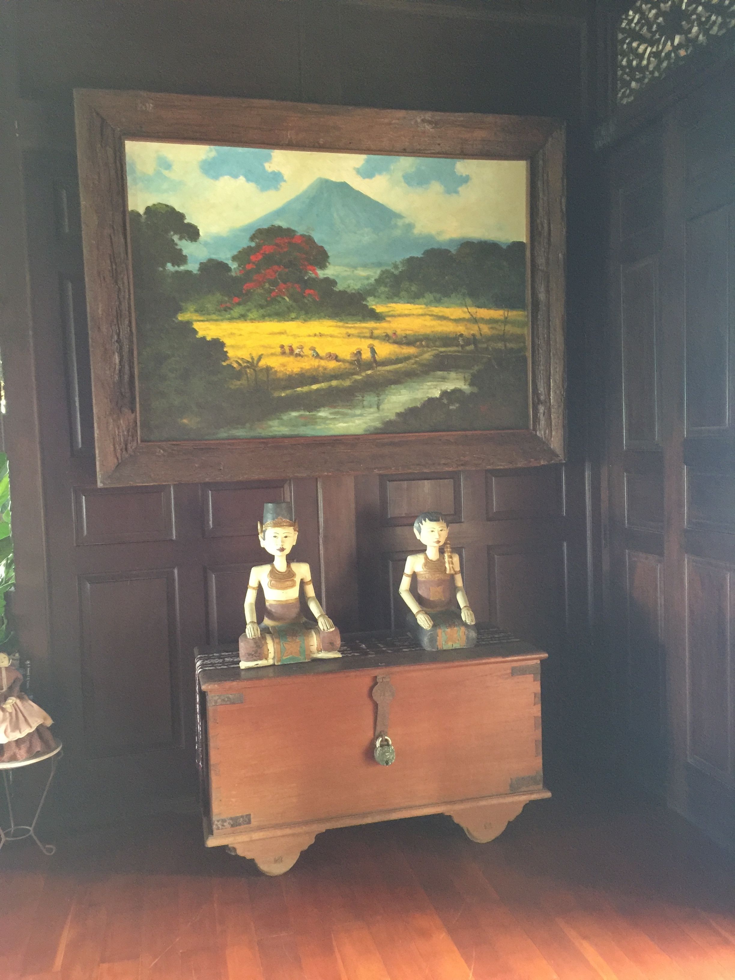A painting of a landscape with a mountain and a river, two wooden sculptures, and a dark wooden cabinet.
