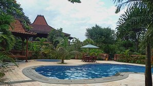A swimming pool with a tiled deck, surrounded by tropical plants and a traditional building with a tiled roof. - Joglo Java (Semarang)