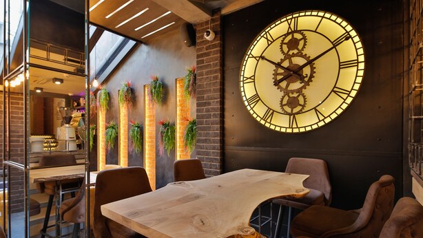 A restaurant interior with a large wall clock, hanging plants, and a unique wooden table centerpiece. - Passpartù (Belgrade)