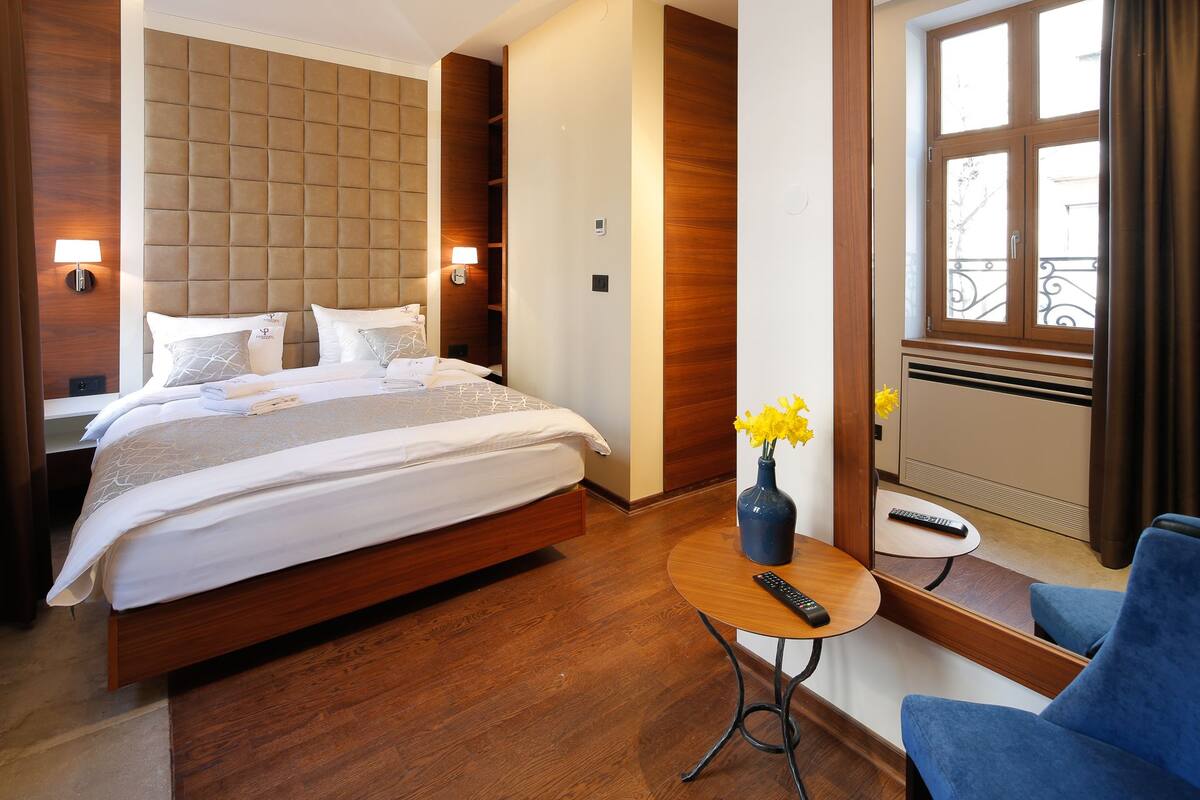 A hotel room with a large bed, a bedside table with a vase of flowers, a round table with a remote control, and a blue chair.