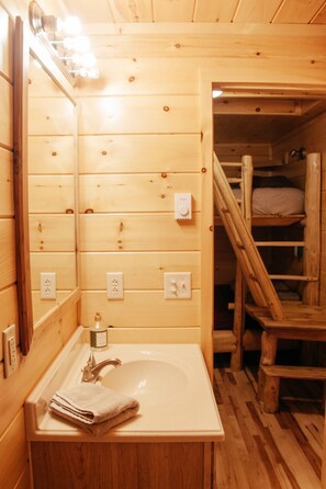 Interior de una cabaña de madera con baño que incluye un lavamanos, espejo y escaleras de madera.