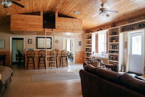 A cozy cabin interior with a dining area, a living room with a leather sofa, wooden ceiling, and a bookshelf filled with books and decorative items. - Wellnesste Lodge and Cabin Rentals (Taberg)