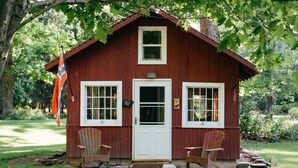 A small red wooden cabin with a white door and two windows, two Adirondack chairs outside, and a flag on the left.