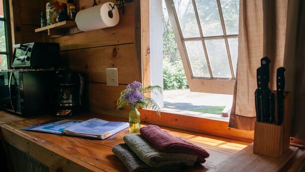 A wooden cabin interior with a wooden desk, a window with curtains, a vase with flowers, and a knife block.
