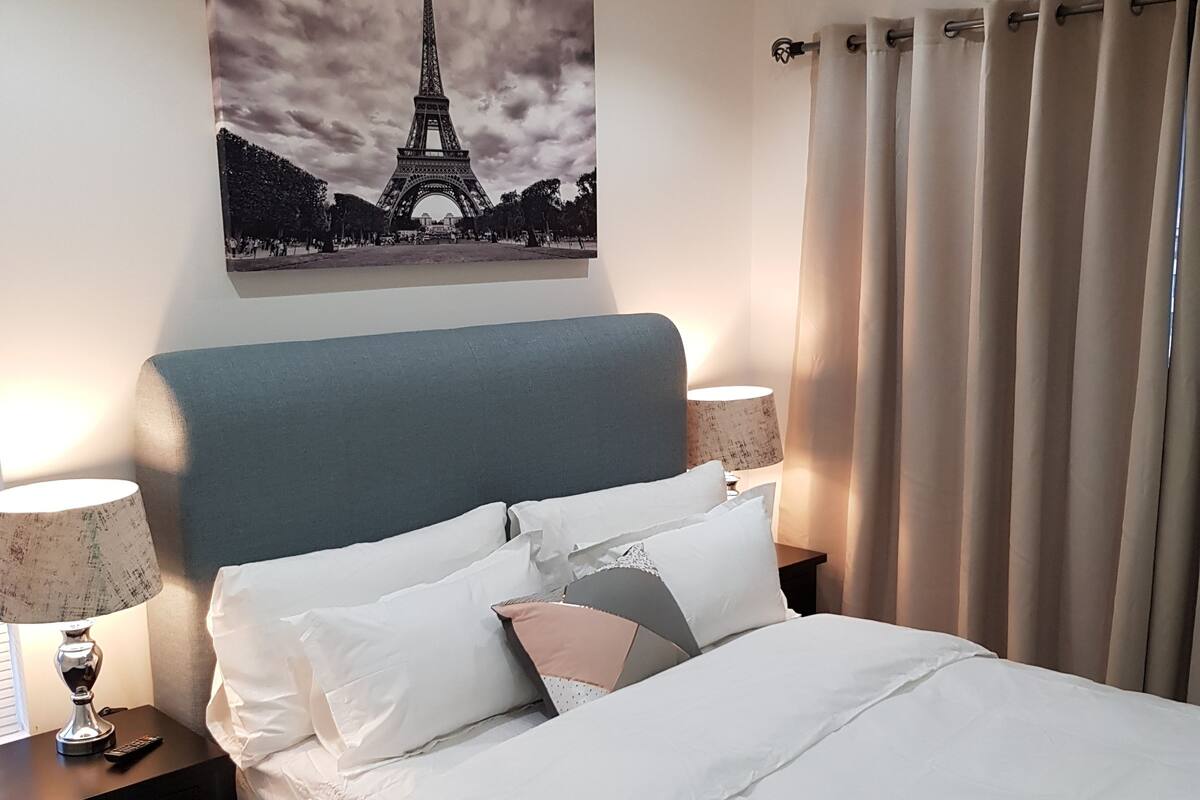 Ein ordentlich bezogenes Bett mit weißen Leinentüchern und mehreren Kissen, eine Nachttischlampe, eine Fernbedienung und ein gerahmtes Bild vom Eiffelturm an der Wand.