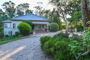 A house with a gravel driveway, a front porch, and a small garden area with various plants and a tree. - Chatelaine Valley Views (Katoomba)