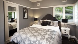 Un dormitorio con cama, mesitas de noche, ventana con persianas y puerta que da a otra habitación.