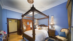 Uma cama de dossel com estrutura de madeira e cortina, um criado-mudo de madeira com uma luminária, uma cadeira com almofada e um espelho grande na parede.