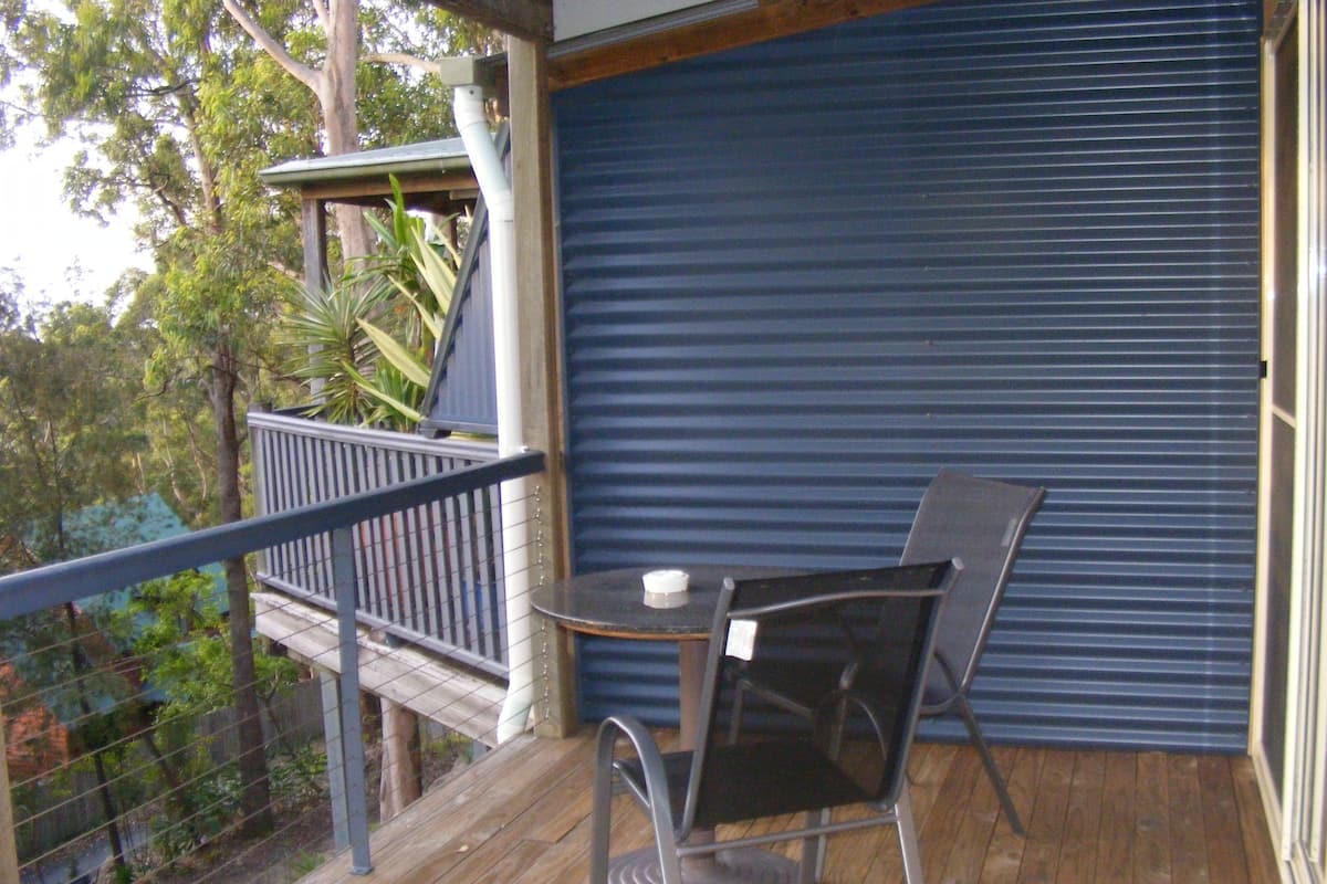A balcony with a table and chairs, a closed roller shutter, and a view of trees.