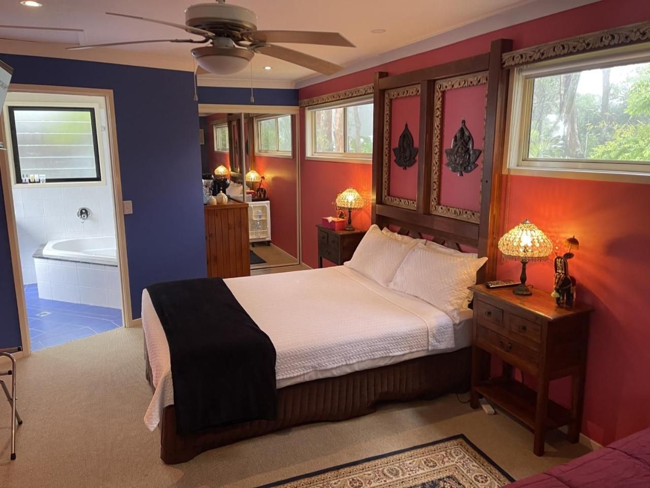 A bedroom with a bed, bedside lamps, a nightstand, a ceiling fan, and a view of a bathroom with a bathtub.