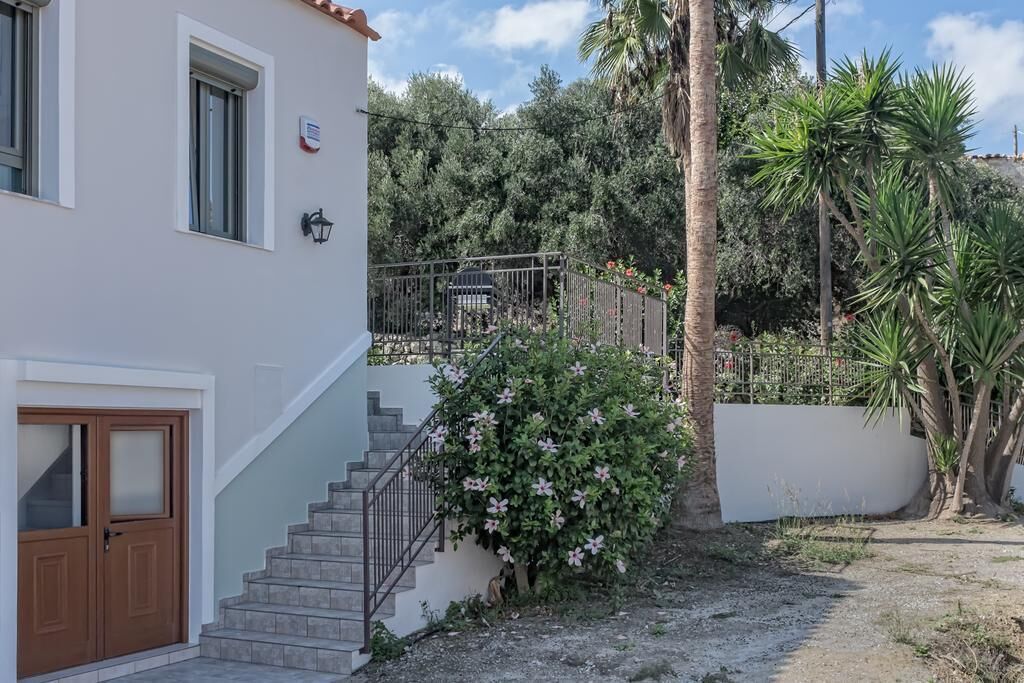 Un edificio a due piani con una scala che conduce a un balcone, immerso nel verde e circondato da un giardino recintato.