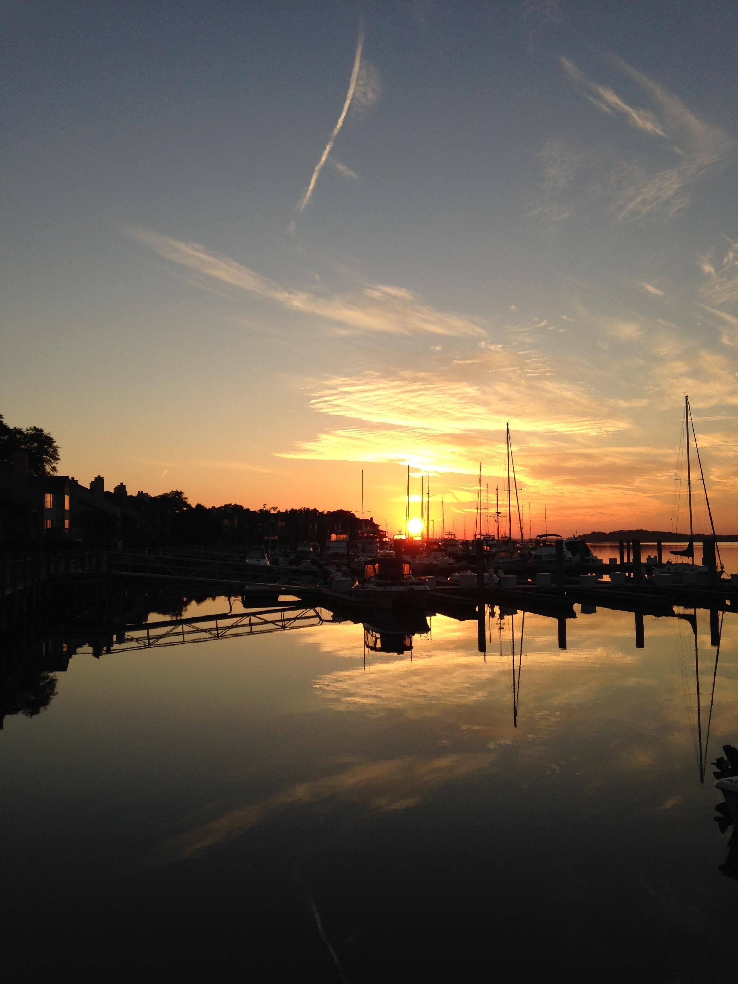 A marina with boats docked, a bridge, and buildings in the background during sunset.