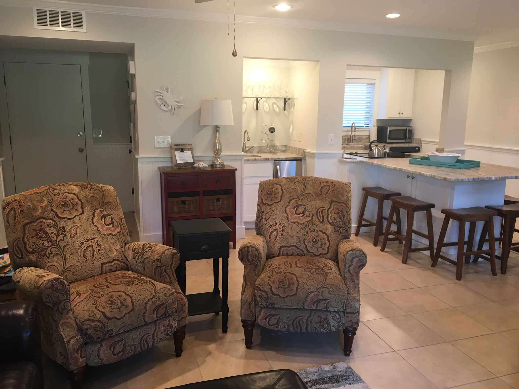 A living room with two patterned armchairs, a small side table, and a sofa. There is a kitchen area with a microwave, sink, and bar stools.