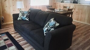 En stue med en mørkegrå sofa, to puder med mønster, trægulv og et vindue.