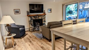 A living room with a stone fireplace, a TV mounted on the wall, a sofa, a dining table, and a view of the outdoors through a large window. - NEW 1 bedroom condo. This is the best location in Ketchum! (Ketchum)