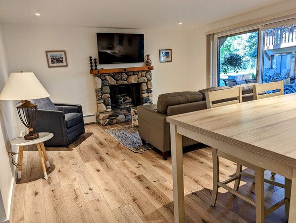 Ein Wohnzimmer mit Kamin aus Stein, einem an der Wand montierten Fernseher, einer Couch, einem Esstisch und Blick ins Freie durch ein großes Fenster.