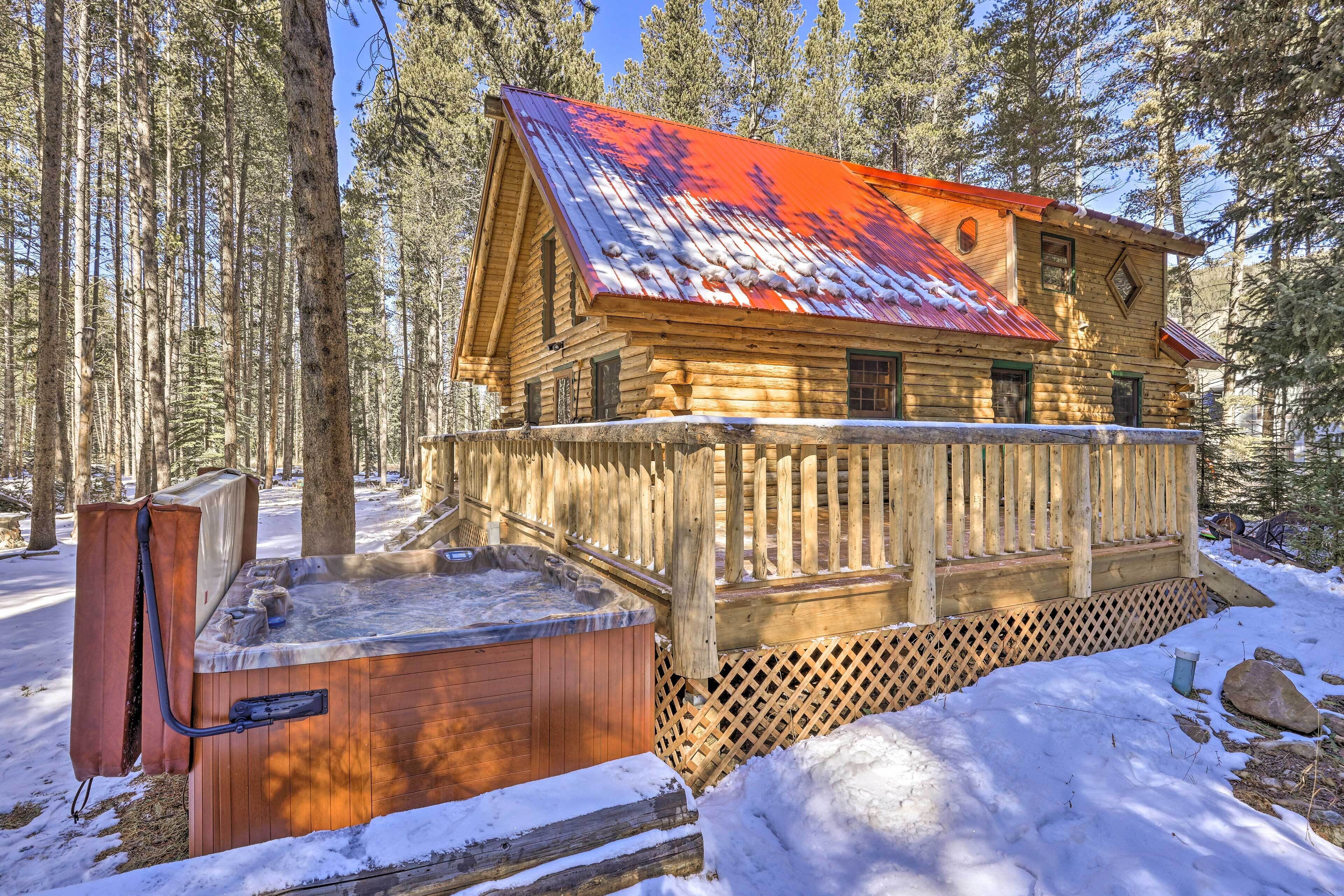 Une cabane en rondins dotée d’un bain à remous et d’une terrasse en bois, entourée d’une clôture en bois et de neige.