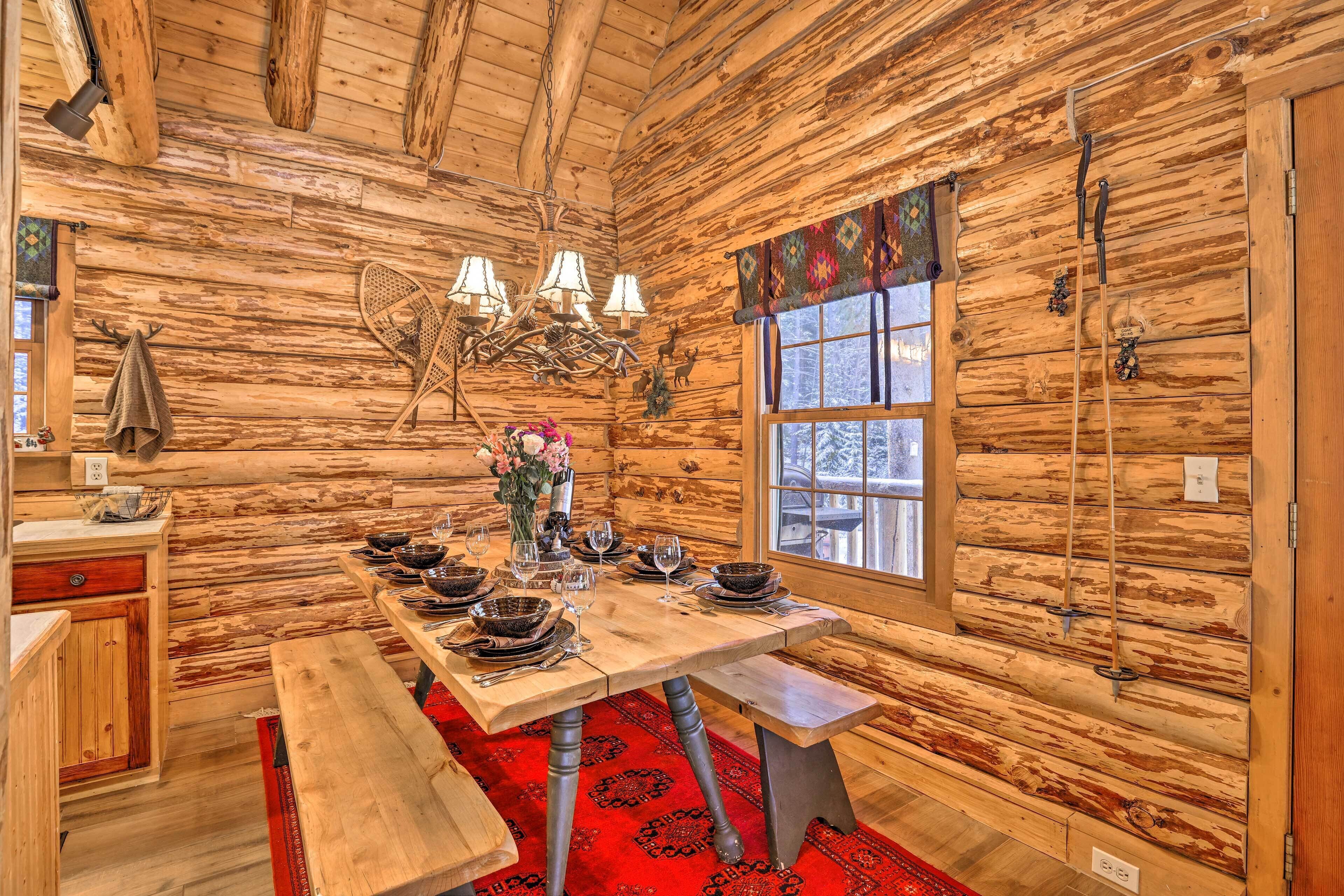 Interior de una cabaña de madera con una mesa de comedor de madera dispuesta para cuatro, un candelabro y una ventana con vistas.