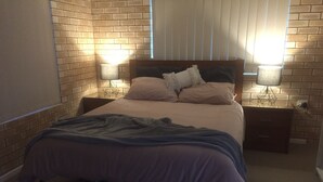 A bedroom with a brick-patterned wall, a bed with a white and blue blanket, two bedside lamps, and a window with blinds. - Holiday at Hillcrest (Scarness)