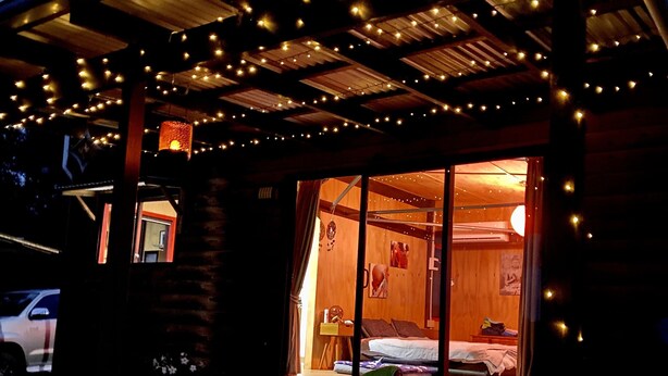 Ein gemütliches Blockhaus mit Veranda, Lichterkette und einem durch das Fenster einsehbaren Bett.