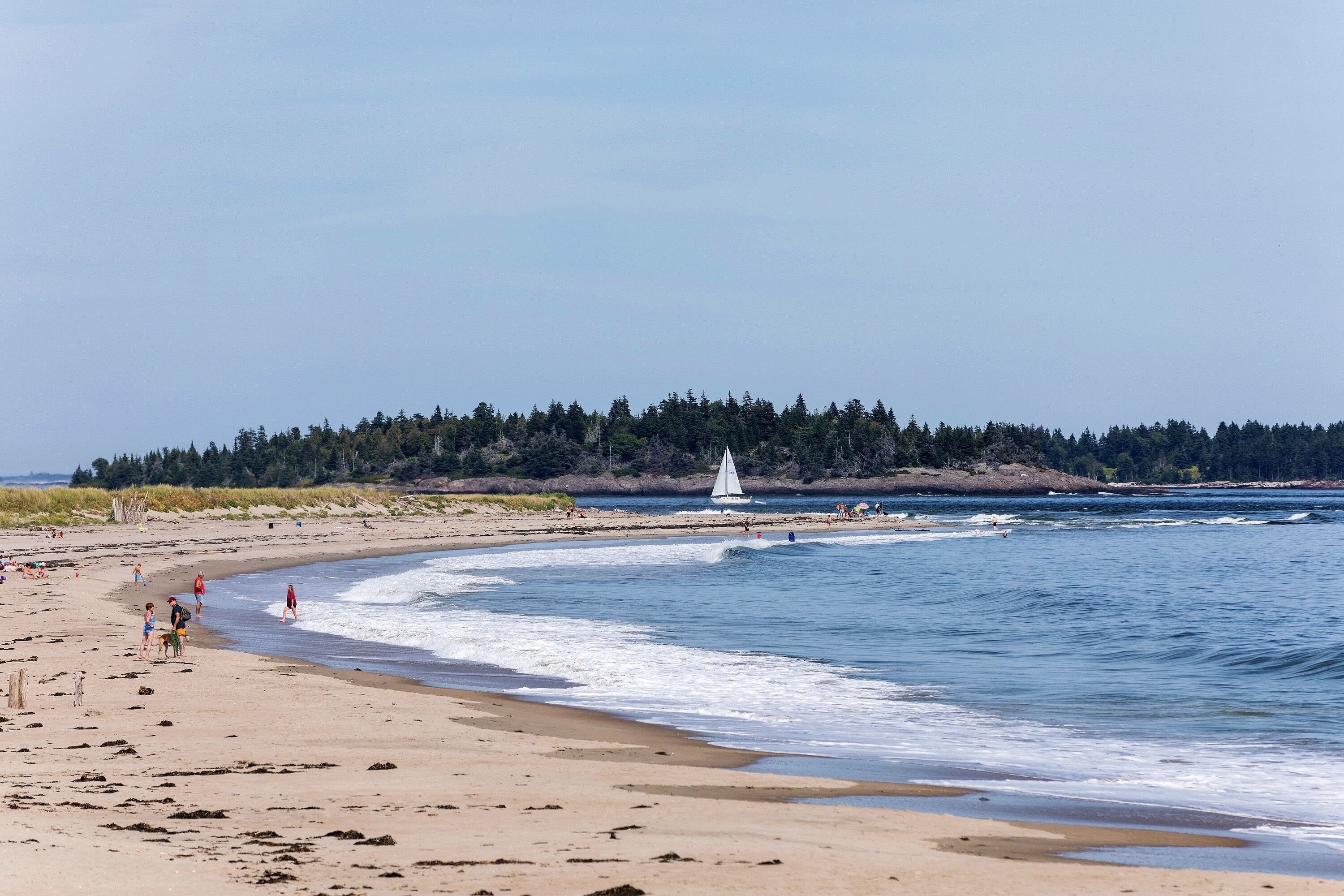 Une plage avec un voilier, des personnes qui se promènent et une zone boisée en arrière-plan.