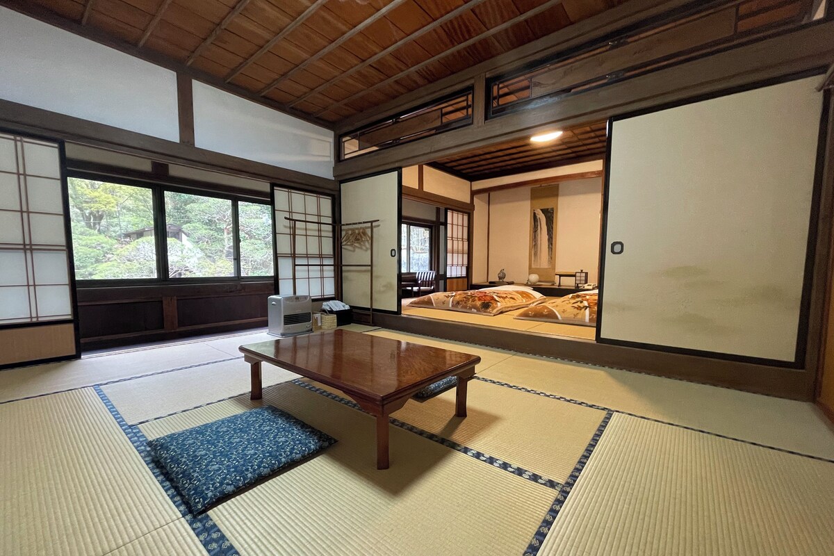 A traditional Japanese room with tatami flooring, a wooden table, sliding doors, and a view of a garden through the windows.