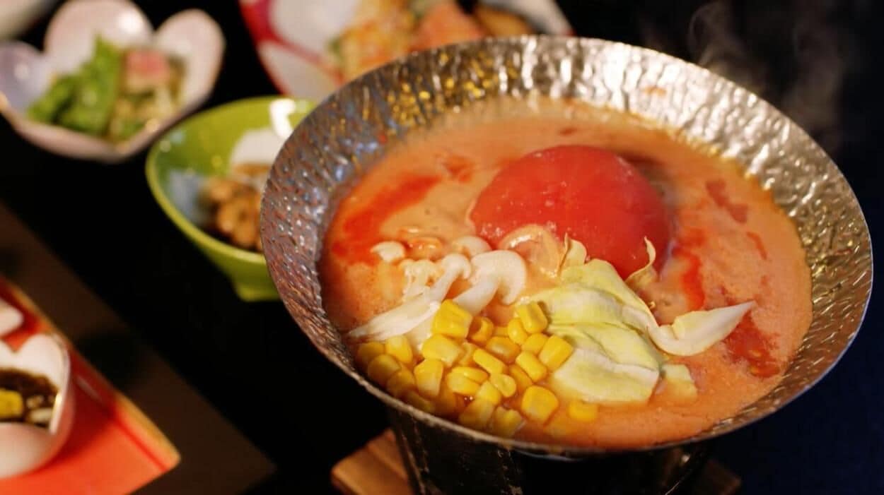 A bowl of soup with corn, cabbage, and a red tomato.