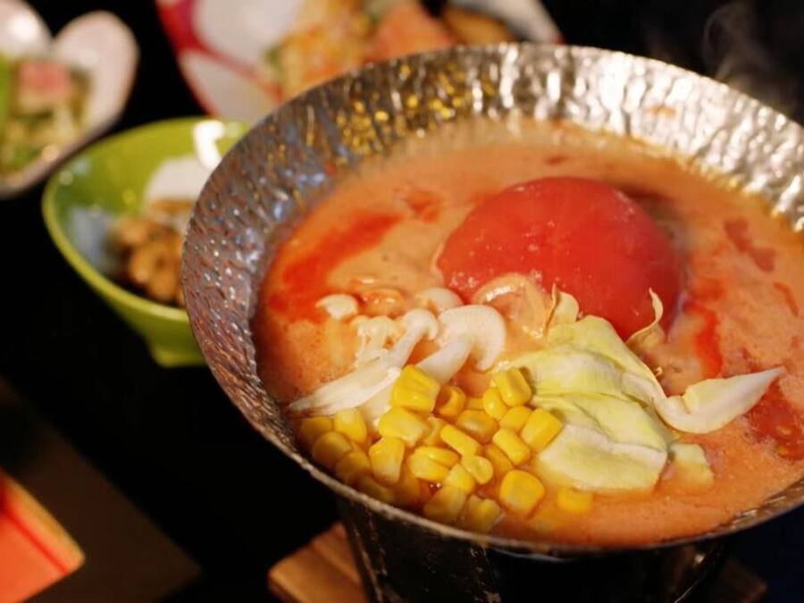 Uma tigela de sopa com milho, repolho e um tomate vermelho.