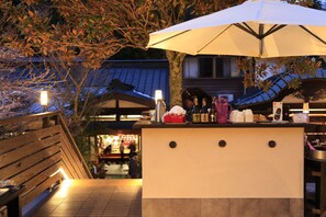 An outdoor seating area with a wooden bench, a table with a white umbrella, and a counter with various items. - Shukubo Kakurinbo (Minobu)