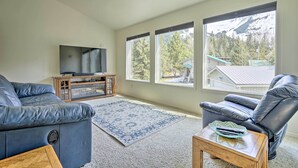 A living room with a TV, two sofas, a coffee table, and a view of mountains through large windows. - Cabin w/ Hot Tub & Views: 1 Mile to Alyeska Resort (Girdwood)
