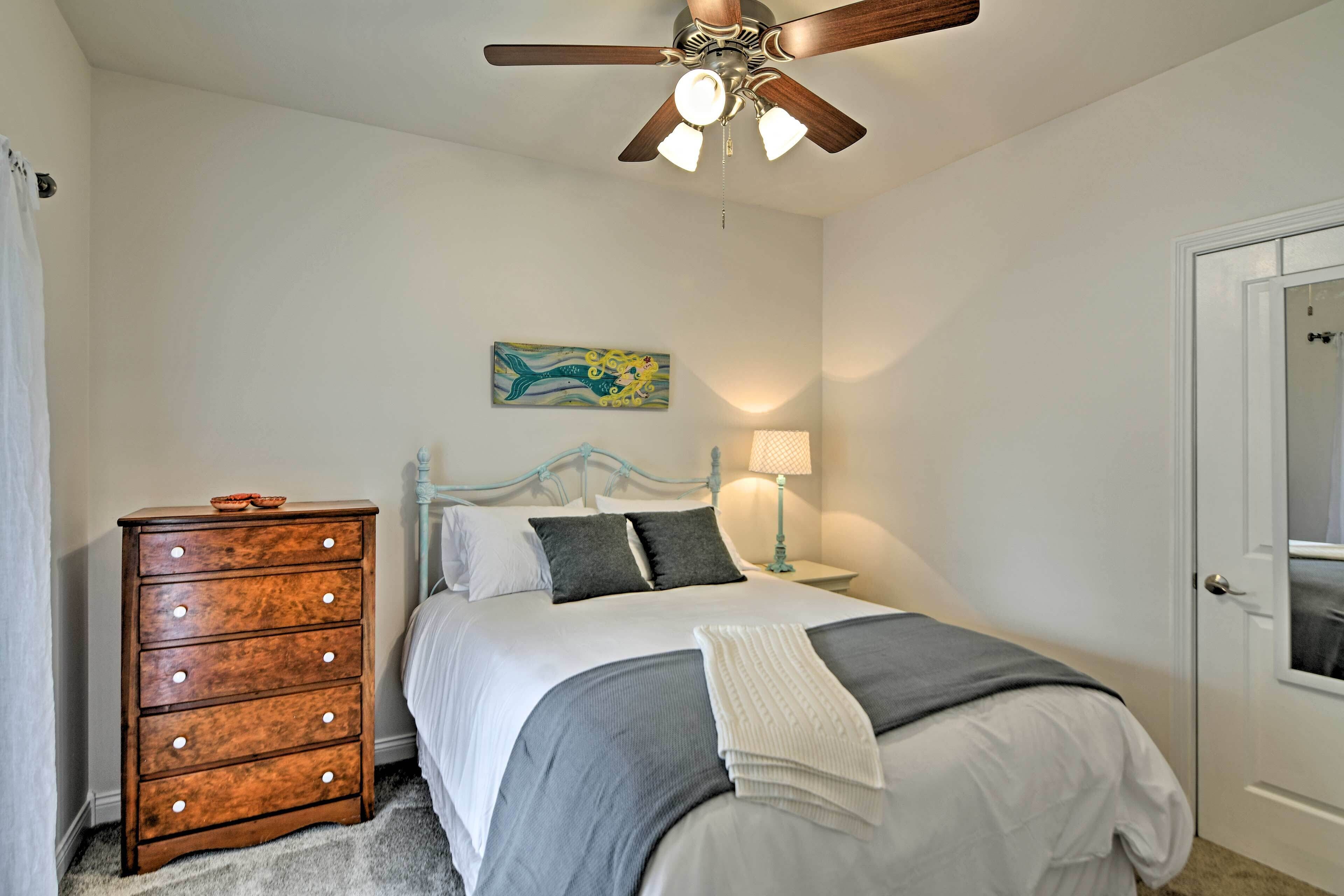 A bedroom with a bed, dresser, ceiling fan, artwork, and a door.