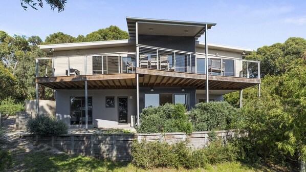 Une maison moderne à deux étages, dotée d’un balcon, de grandes fenêtres et d’un mur de soutènement en pierre.