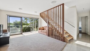 Ein geräumiger Wohnbereich mit einer großen Schiebeglas-Tür, die auf einen Balkon führt, einer Holztreppe und einem Flachbildfernseher auf einem Standfuß.