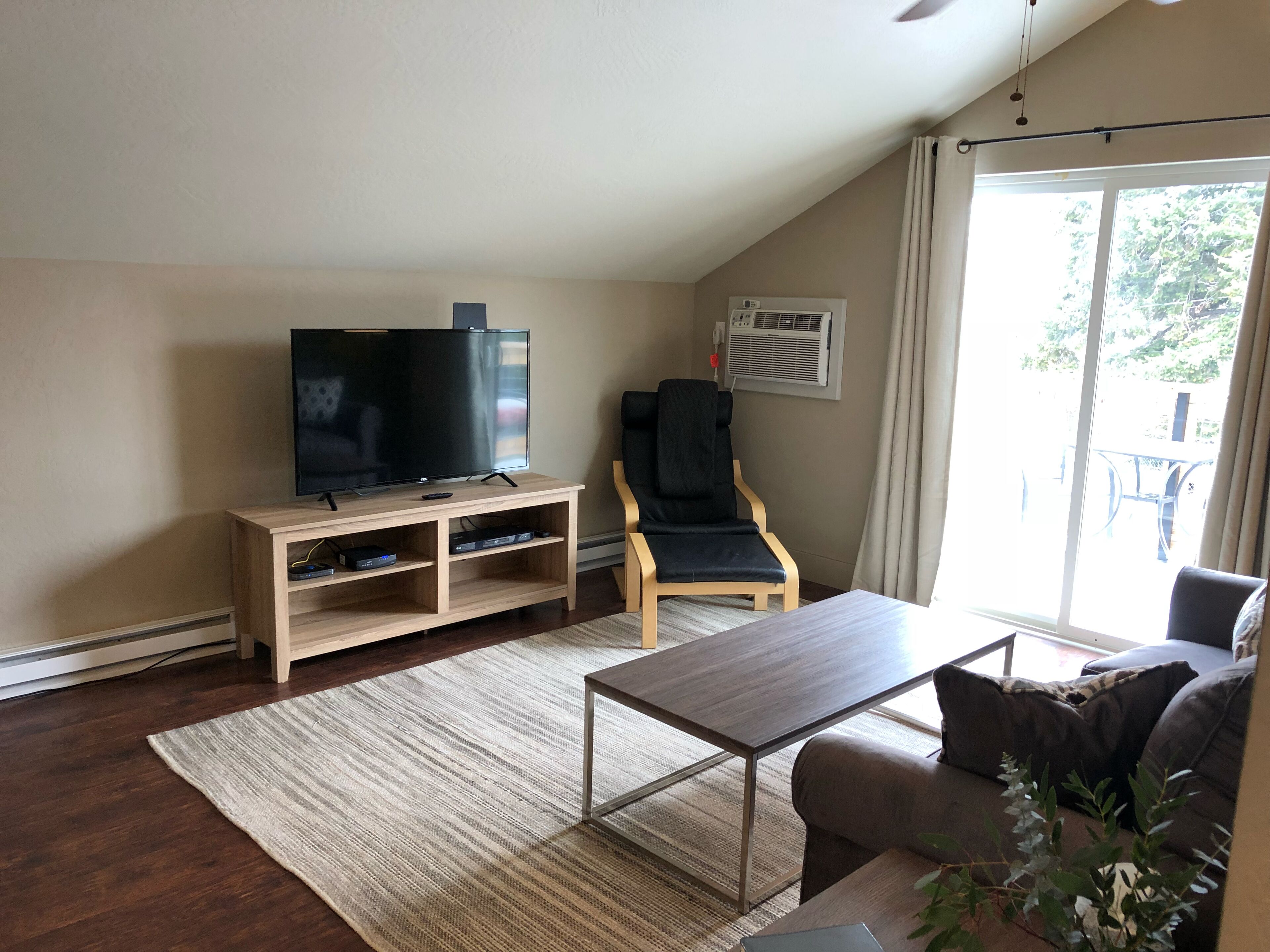 A living room with a flat-screen TV on a wooden stand, a reclining chair, a coffee table, and a large window with curtains.