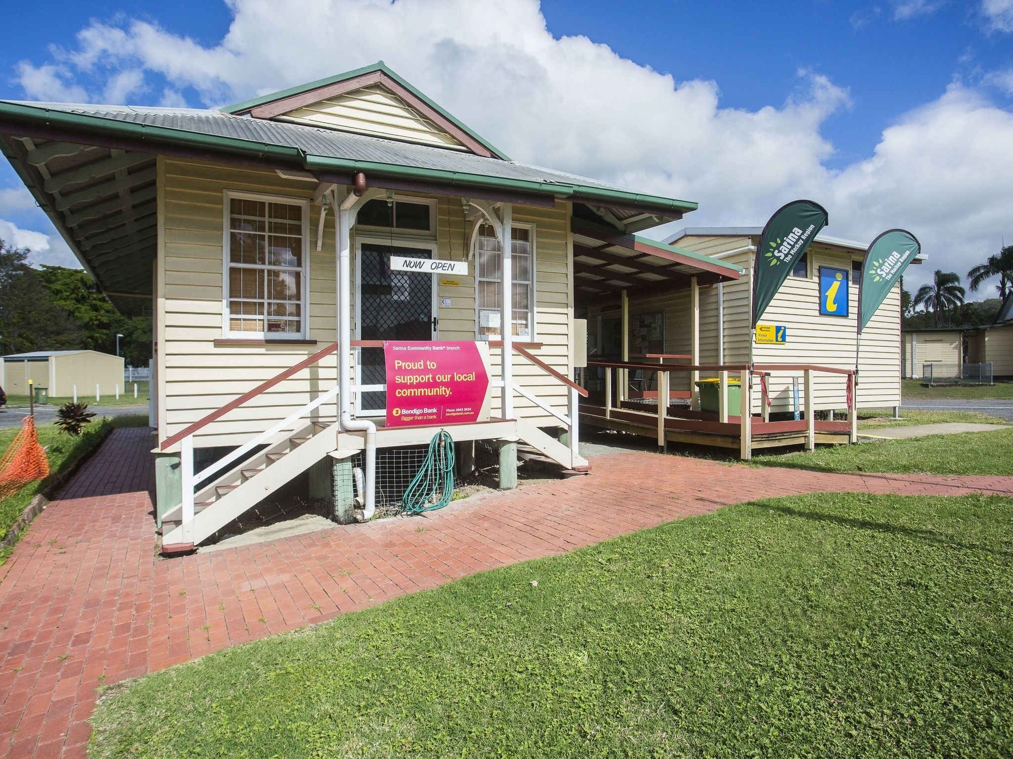 A small, single-story building with a green roof and a red brick pathway leading to it. There is a sign on the building that reads "NOW OPEN" and another sign that says "Proud to support our local community."