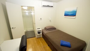 A small, single-room hotel room with a bed, a desk, a chair, a small bedside table, a wall-mounted air conditioner, and a painting. - The Three Ways Alligator Creek (Alligator Creek)