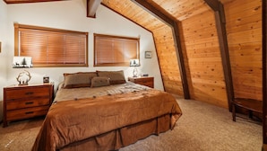 Un dormitorio con techo inclinado, paredes de madera, una cama con colcha marrón, dos mesitas de noche con lámparas y dos ventanas con persianas.