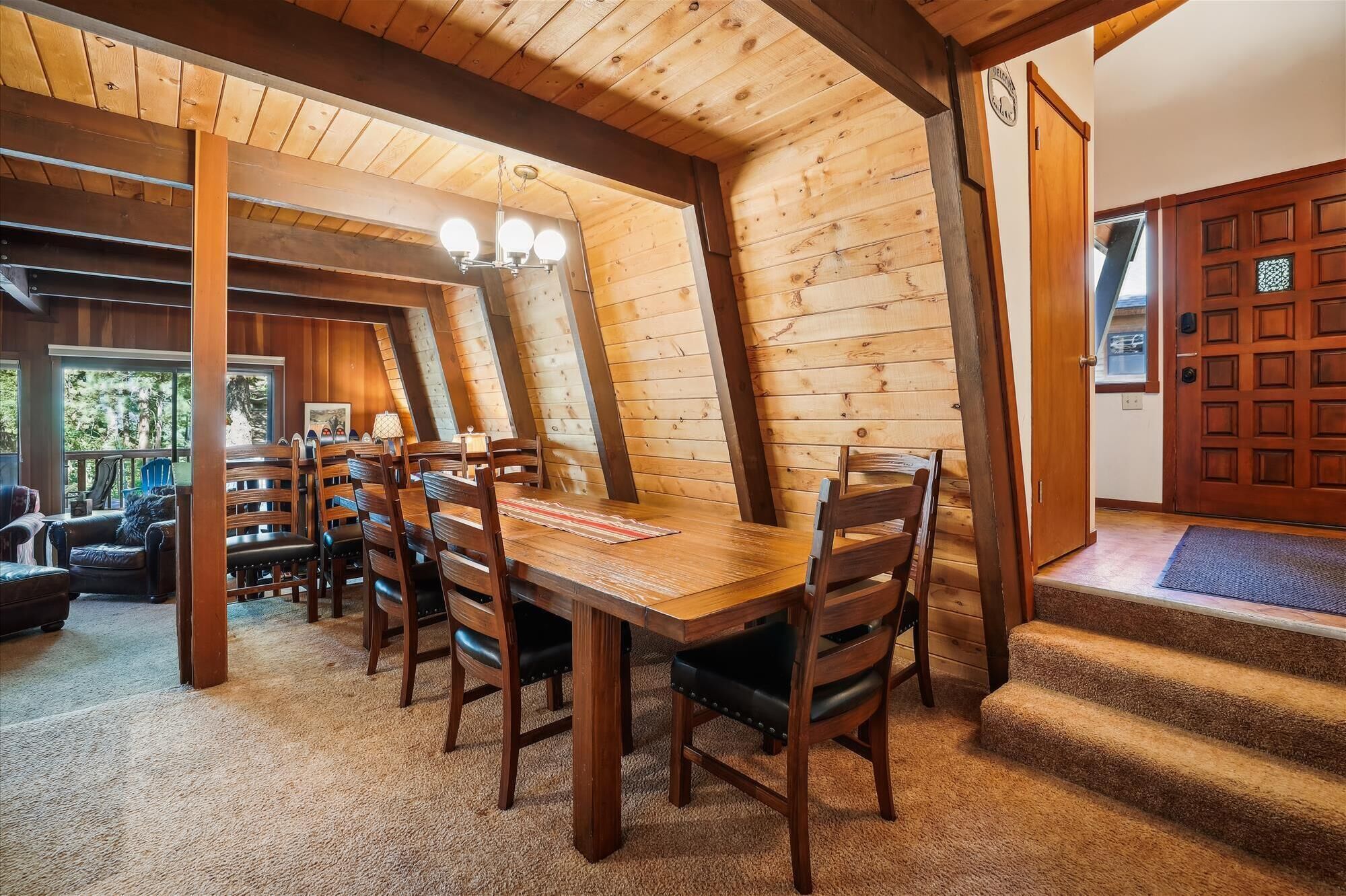 A dining area with a wooden table and chairs, a chandelier, and a view of the outdoors through large windows.