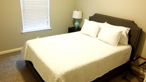 Una cama bien tendida con una colcha blanca y dos almohadas, una mesita de noche con una lámpara azul turquesa y una ventana con persianas.