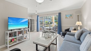 Ein Wohnzimmer mit Flachbildfernseher, einem weißen Couchtisch, einem grauen Sofa, einem Esstisch mit Stühlen und einem Deckenventilator.